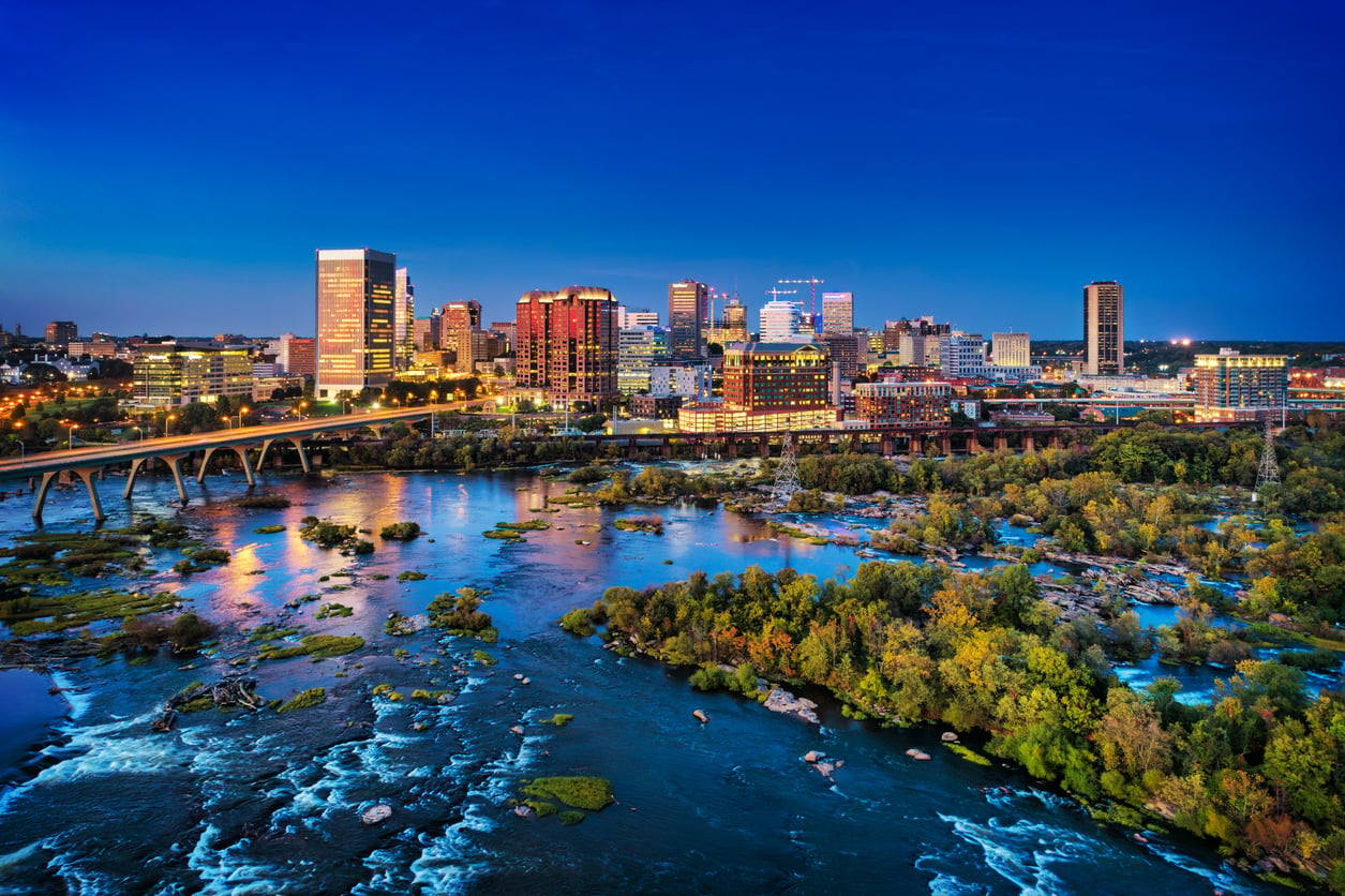 City skyline at dusk with tall buildings lit up, a travel-friendly river with small rapids and rocks in the foreground, and a bridge crossing the water. Trees and greenery line the riverbanks under a deep blue sky.