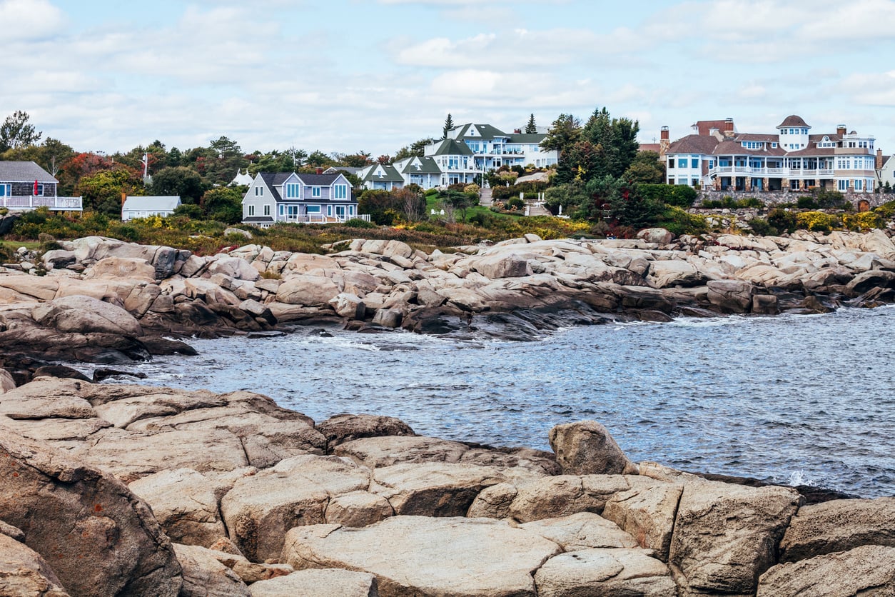 Rocky shoreline with waves, several houses, and trees in the background under a partly cloudy East Coast sky.