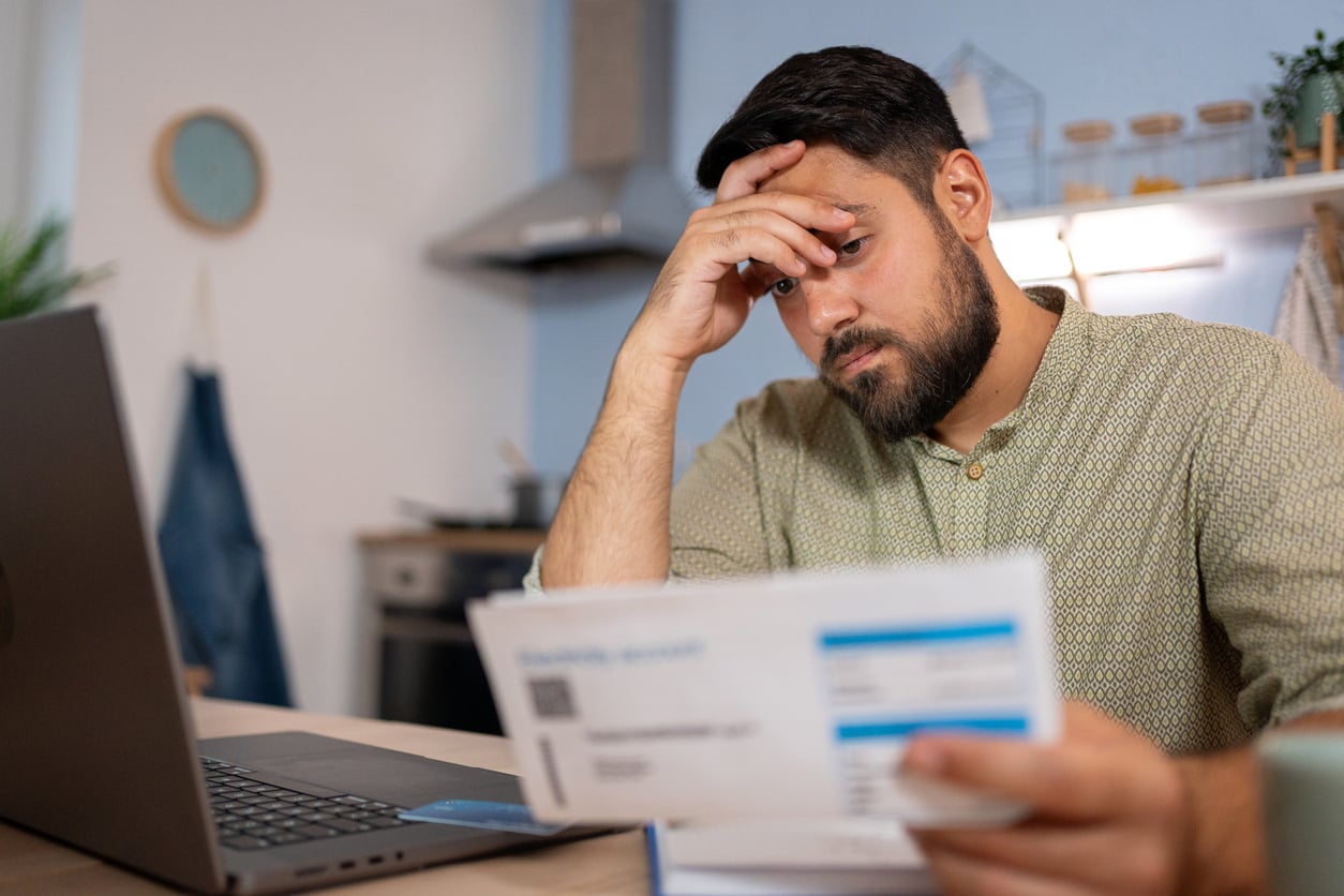 A man sits at a table with a laptop, holding and looking worriedly at a bill or financial document in his hand, in a home kitchen setting, thinking about developing better money saving habits.