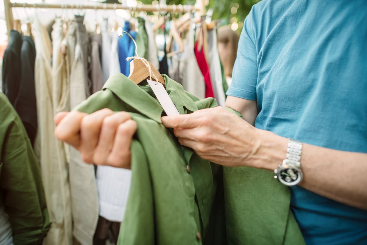 A person in a blue shirt examines a green button-up shirt on a hanger at a clothing rack, possibly at a market or store, showcasing money saving habits while shopping.
