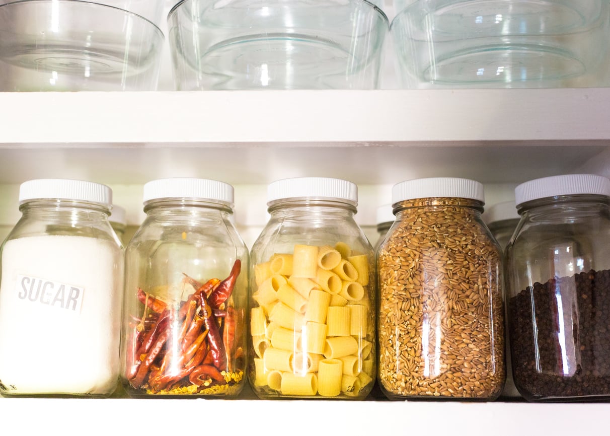 Five glass jars on a shelf, filled with sugar, dried chilies, pasta, grains, and black peppercorns—each reflecting thoughtful grocery shopping habits. Empty glass bowls are on the shelf above. Bright, natural lighting fills the scene.