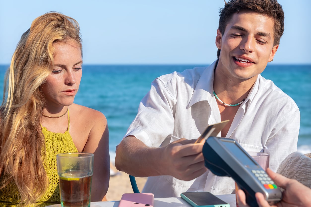 A man pays with a card using a wireless terminal at an outdoor seaside café, enjoying beach vacation savings while a woman with long blonde hair sits beside him, both with drinks on the table.