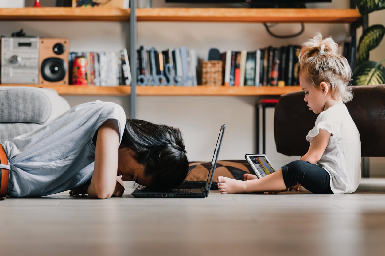 A woman rests her head on a laptop keyboard, appearing tired, while a young child sits nearby on the floor using a tablet—a quiet snapshot reflecting how families in America are having fewer children in cozy, book-filled living rooms.