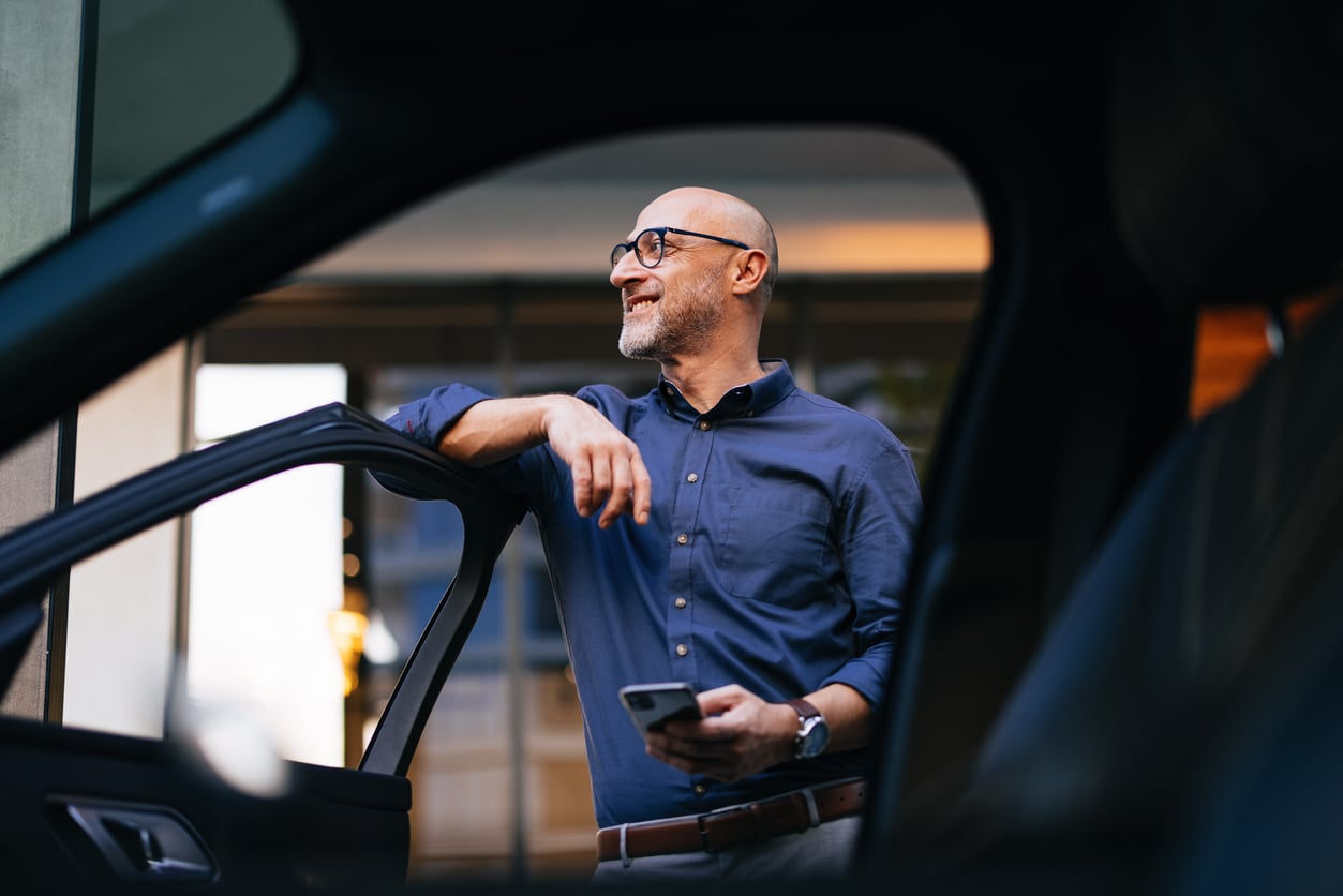 A smiling man wearing glasses and a blue shirt stands outside an open car door, holding a smartphone—perhaps searching for car maintenance hacks—as he rests his arm on the car and looks off into the distance.