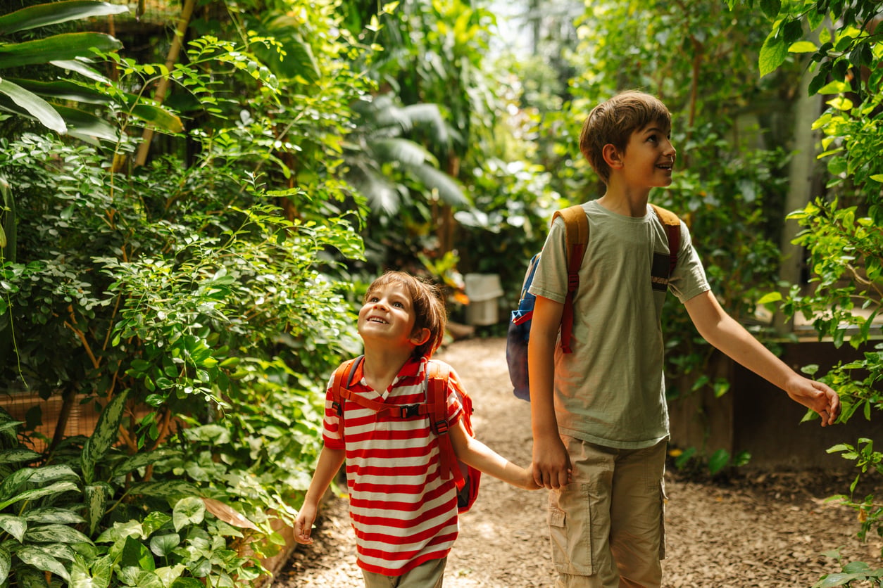 Two children with backpacks walk hand in hand along a lush, green garden path. One child looks up smiling, while the other looks ahead, surrounded by vibrant foliage—a scene that captures the kind of adventures that make memberships worth paying for.