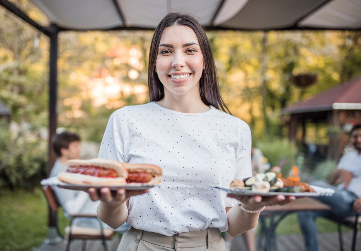 A woman in a white shirt smiles while holding plates of food, including a hot dog, outdoors at a gathering with blurred people and greenery in the background.