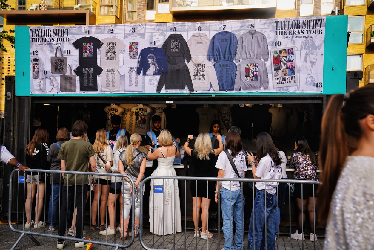 Fans line up at a Taylor Swift Eras Tour merchandise stand, with shirts and hoodies displayed above the booth—unlike products people no longer buy, these items remain highly sought-after. The scene is outdoors and fenced with metal barricades.