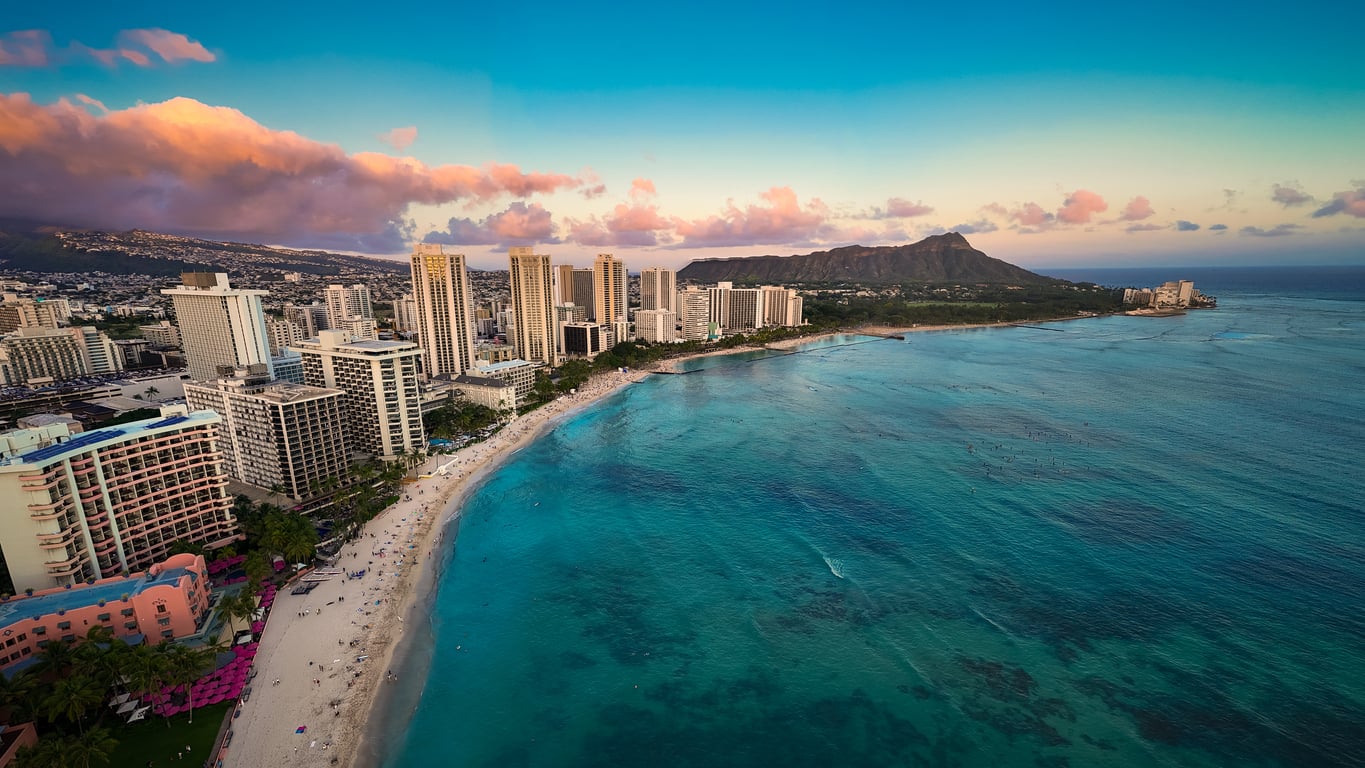 Aerial view of a city skyline with tall buildings along a sandy beach, turquoise ocean, and a distant green volcanic mountain under a colorful sunset sky—a stunning destination where vacation costs may be well worth the unforgettable experience.
