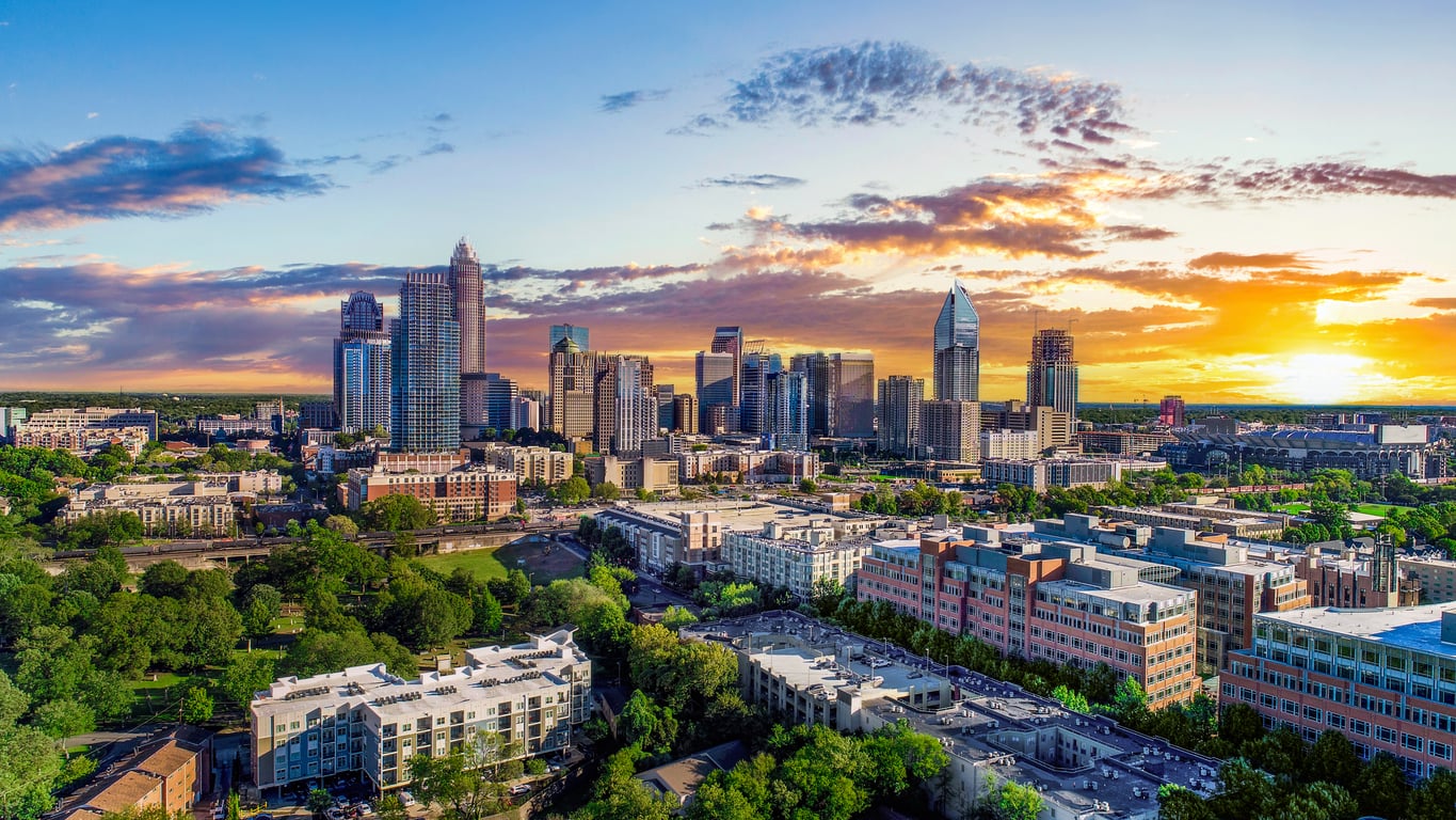 Aerial view of a travel-friendly city skyline at sunset with tall buildings, green trees, and a partly cloudy sky, sunlight illuminating the scene.