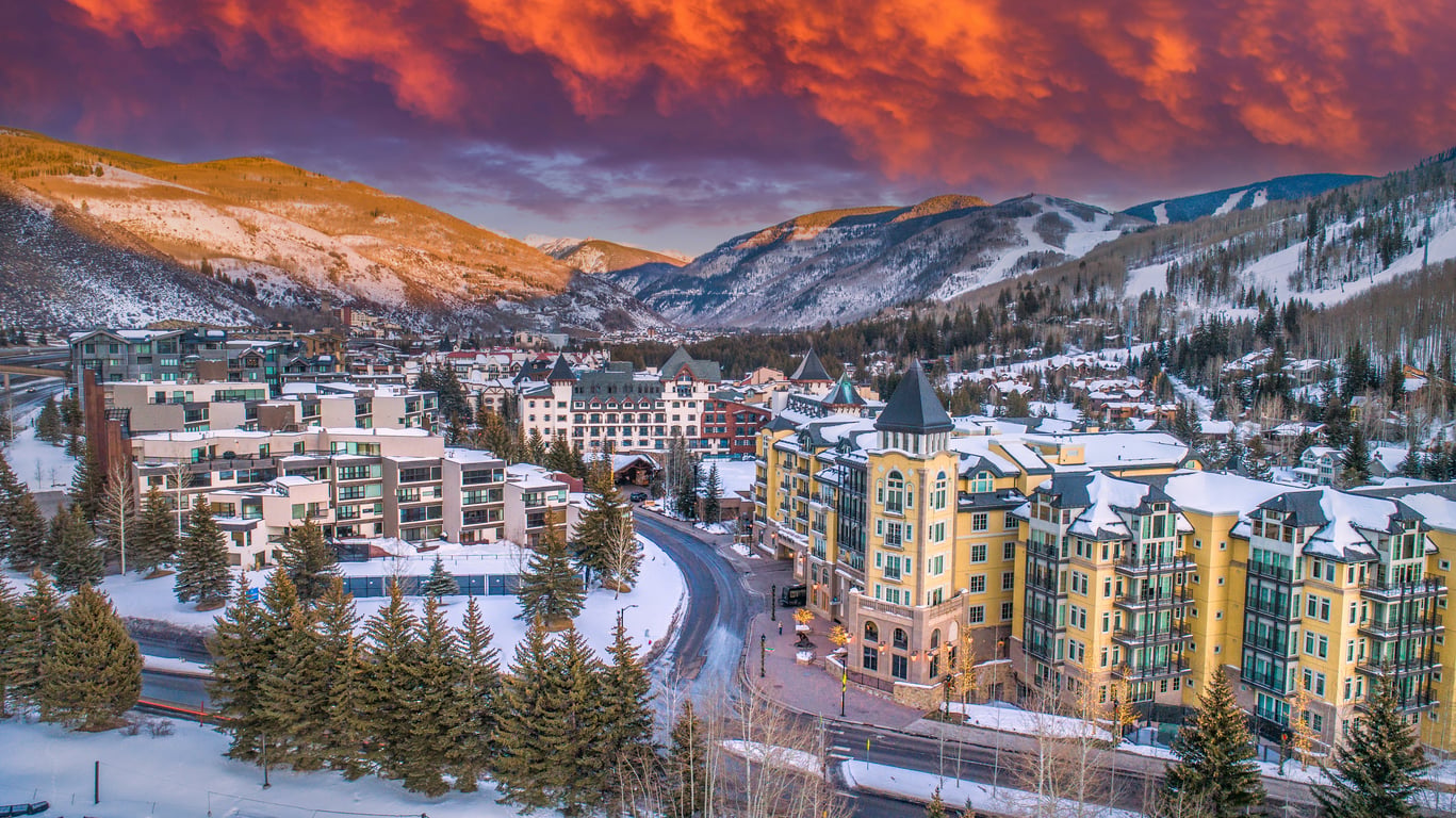 Snow-covered mountain town with colorful buildings and evergreen trees, nestled in a valley at sunset. The sky above glows with dramatic orange and purple clouds, illuminating the surrounding peaks and creating a warm, scenic atmosphere—making it one of the most captivating travel destinations for those seeking breathtaking views and cozy charm.