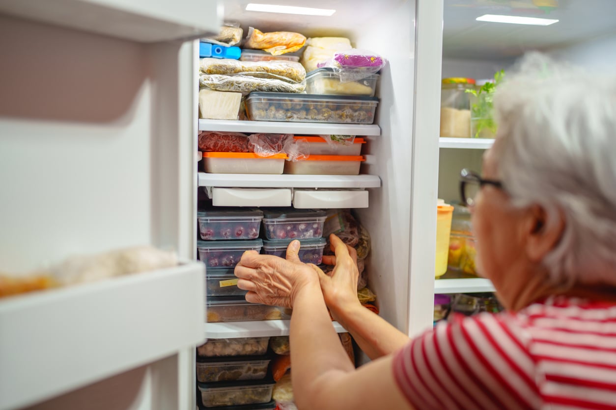 An elderly person wearing glasses and a red-striped shirt arranges plastic containers and bags of food inside a packed freezer, reflecting thoughtful grocery shopping habits.