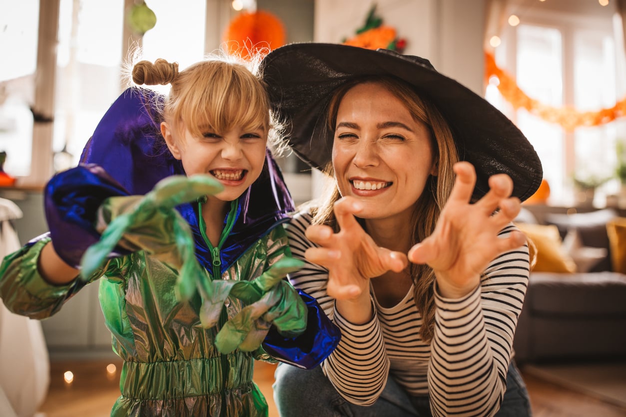 A woman in a witch hat and a child in a shiny costume smile and pose with clawed hands, pretending to be scary for Halloween in a decorated room, enjoying fun Halloween activities together.