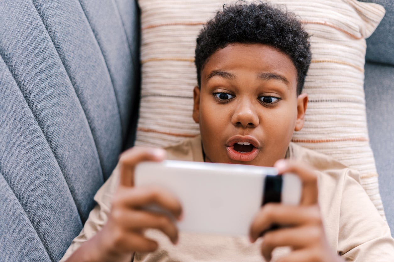 A boy lies on a sofa, looking surprised and excited while holding a smartphone in both hands, possibly playing a game or watching something.
