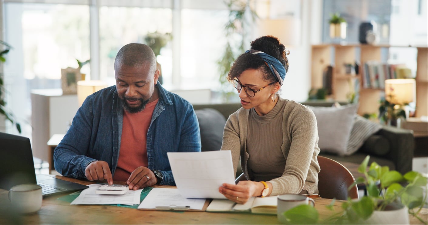 A man and a woman sit together at a table reviewing documents about the benefits of renting. The man uses a calculator while the woman holds a piece of paper. They appear focused, surrounded by plants and office supplies in a bright room.