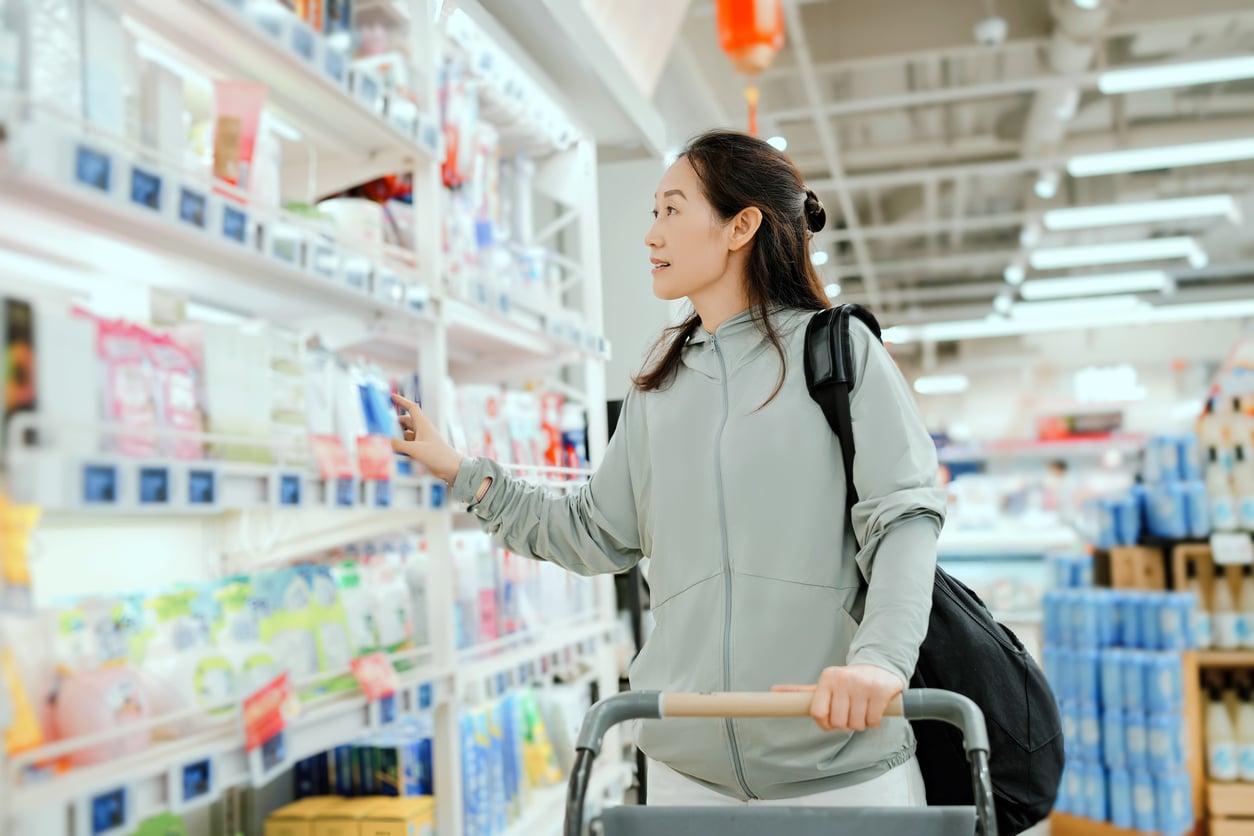 A woman in a light jacket shops in a brightly lit store, pushing a shopping cart and reaching for an item on a shelf stocked with various products—perhaps gathering essentials before setting off on one of her upcoming vacation swaps.
