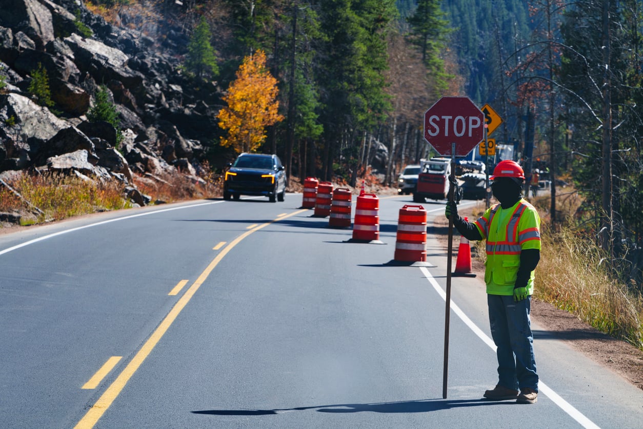 A construction worker in a neon jacket and hard hat holds a stop sign on a two-lane road with orange barrels and cars waiting, surrounded by trees and rocky hills—an unexpected pause that reminds travelers of the importance of having a reliable road trip checklist.