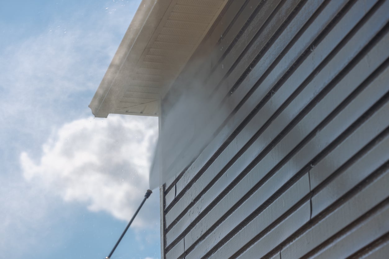 A person uses a pressure washer to clean the exterior vinyl siding of a house on a sunny day, with water spray visible against the building and a blue sky with clouds in the background.