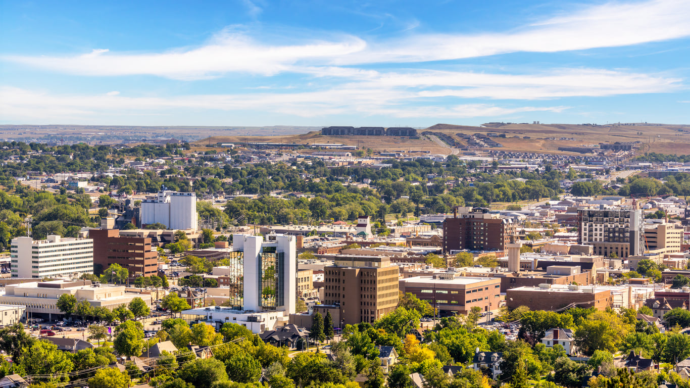 Aerial view of a mid-sized city with modern and older buildings, surrounded by green trees—one of the 20 travel destinations renowned for its stunning mix of architecture. Hills and a blue sky with wispy clouds are visible in the background.