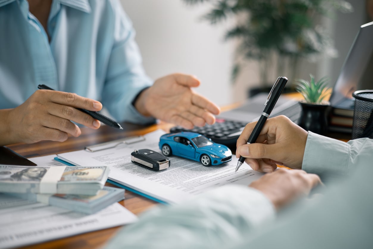 Two people sit at a desk discussing a car purchase contract. Toy cars, a car key, cash, and documents—possibly detailing monthly bills—are on the table. One person gestures while the other signs papers. A calculator and plant are in the background.