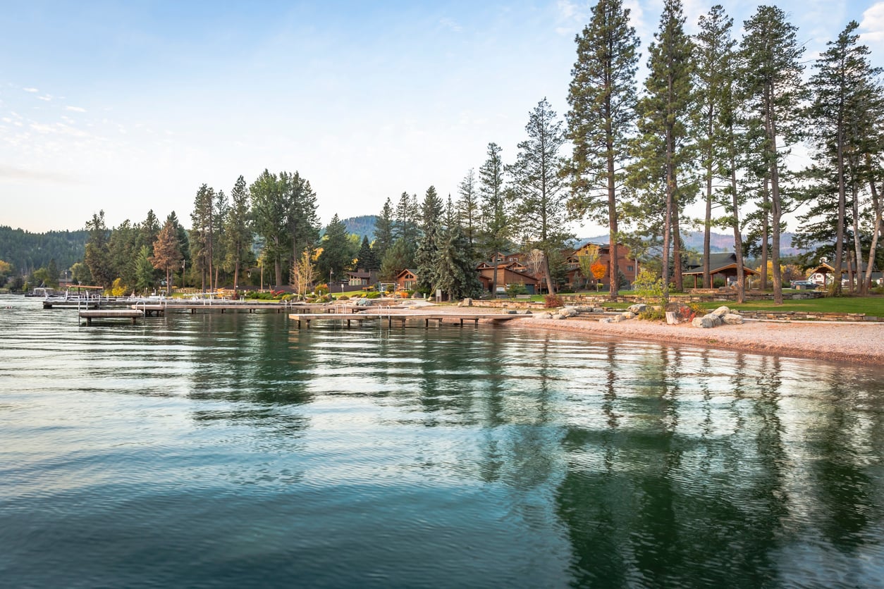 Lakeside view with clear water, wooden docks, and cabins surrounded by tall pine trees. Mountains and a partly cloudy sky are visible in the distance.