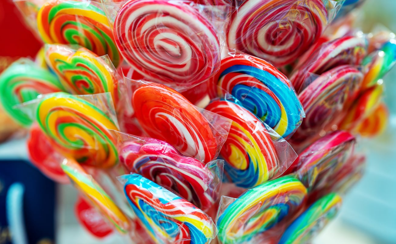 A close-up of colorful, swirled lollipops wrapped in plastic, grouped together. The lollipops have bright red, yellow, blue, green, and pink patterns.