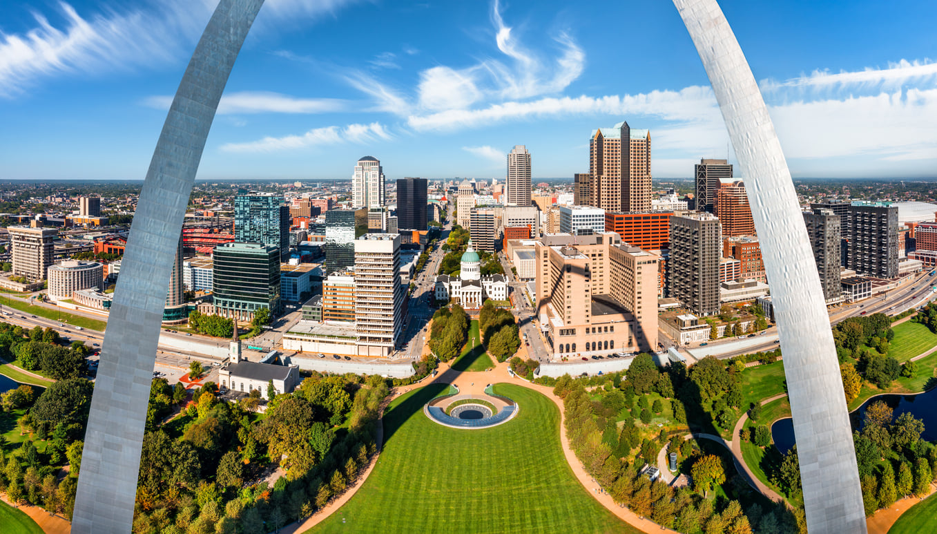Panoramic view from the top of the Gateway Arch in St. Louis, Missouri—one of the iconic landmarks found in big cities in America. The frame shows the sweeping metal legs of the Arch on either side, overlooking the city skyline, the Old Courthouse, and green park grounds under a bright blue sky.