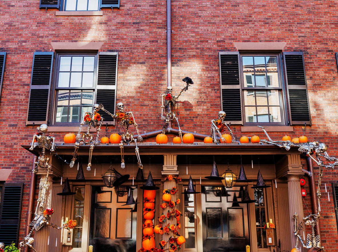 Halloween decorations on a brick building: skeletons sit and climb on the roof, surrounded by pumpkins and orange lights, creating a festive, spooky display above the entrance—just the kind of scene you’d expect to find in one of the best trick or treat towns in America.