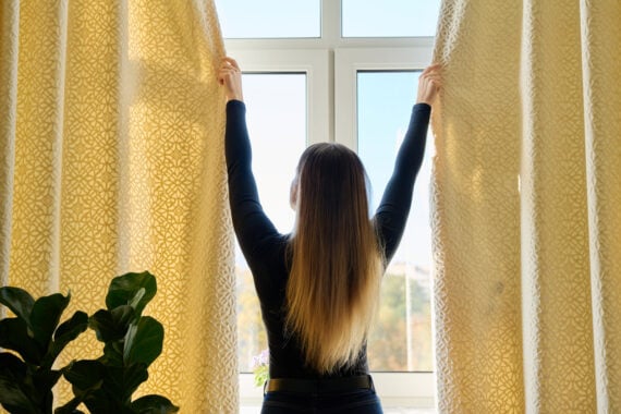 A person with long hair, seen from behind, opens yellow patterned curtains to let sunlight into a bright room. A green leafy plant is visible on the left side of the image.