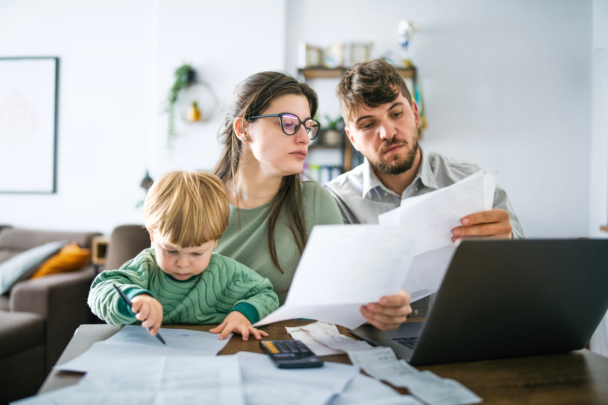 A couple sits at a table reviewing papers and using a laptop, while their young child draws with a pencil—an everyday scene reflecting how families in America are having fewer children. Bills, papers, and a calculator are spread across the table in a home setting.