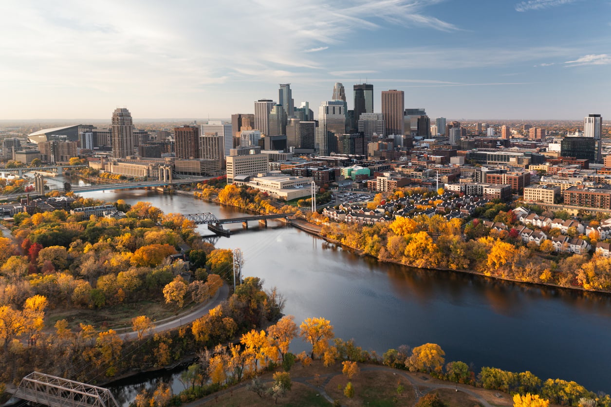 Aerial view of a travel-friendly city skyline with tall buildings, a river with bridges, and trees with bright autumn foliage under a partly cloudy sky.