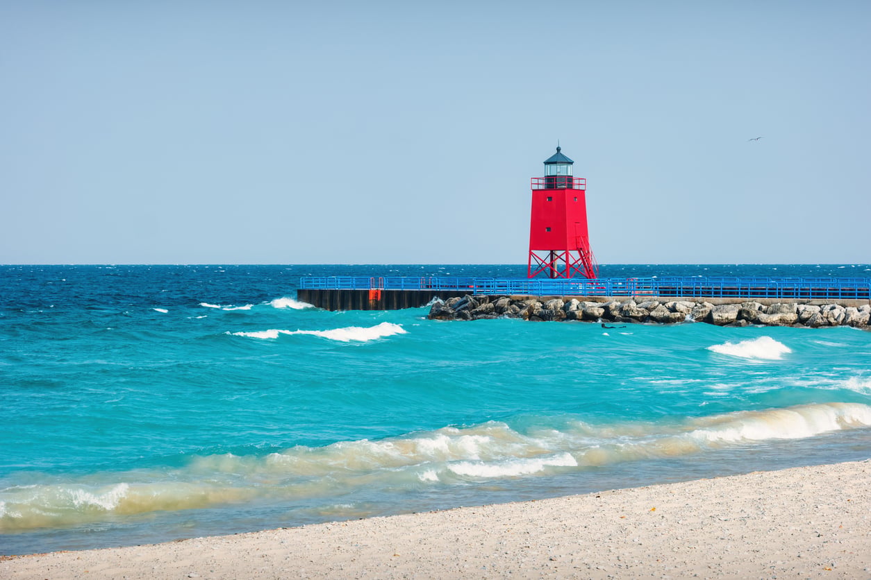 A bright red lighthouse stands on a rocky pier by turquoise waves under a clear blue sky, with sandy beach in the foreground—capturing the timeless charm often found in classic beach towns.