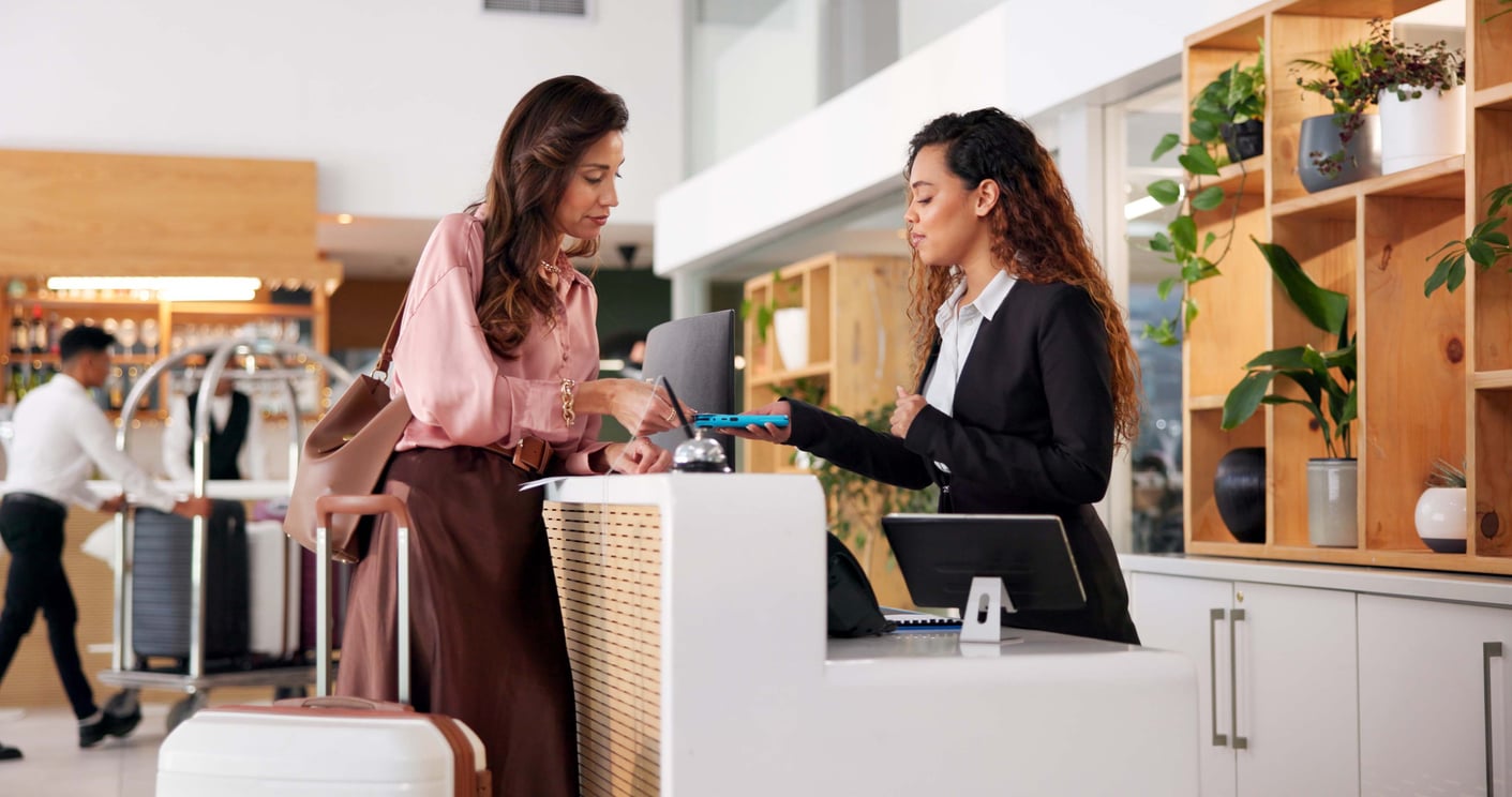 A woman checks in at a hotel reception desk, speaking with a receptionist in a suit. The guest has a suitcase and handbag, while the modern reception area—with wooden shelves and plants—reflects recent hotel changes.
