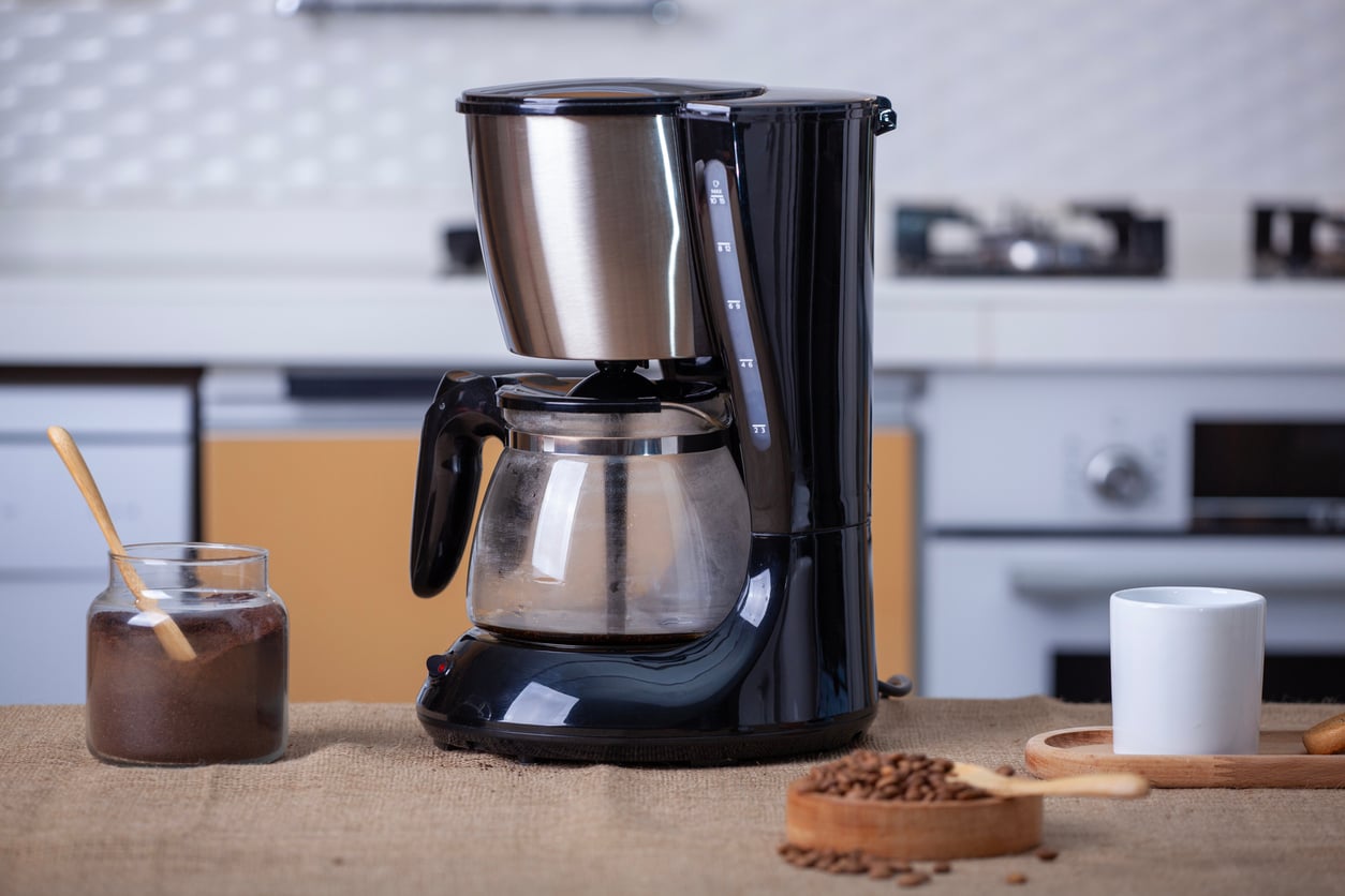 A black drip coffee maker sits on a kitchen counter next to a jar of ground coffee with a spoon, a white coffee mug, a wooden tray, and scattered coffee beans—an inviting display of essential kitchen items. Kitchen appliances are visible in the background.
