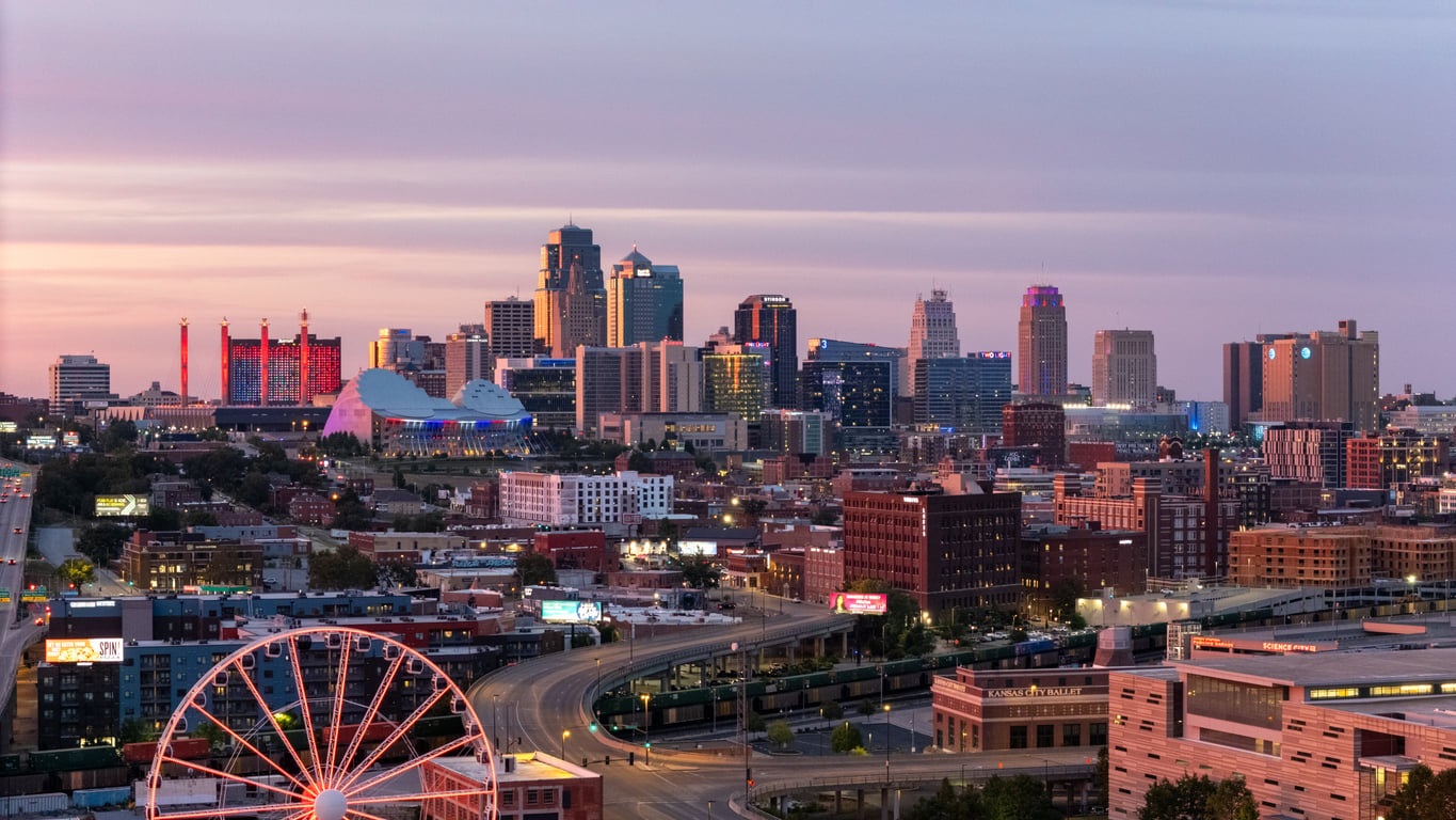 Kansas City skyline at sunset with tall buildings, a ferris wheel in the foreground, and colorful lights illuminating some structures—an urban scene that easily earns its place among the top 20 travel destinations for vibrant cityscapes.