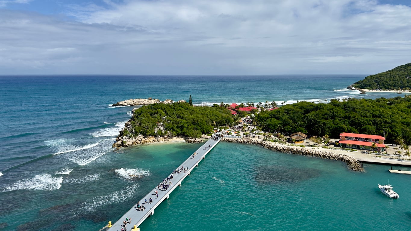 Aerial view of a pier stretching over turquoise water toward a lush, green coastal area with buildings and people, bordered by ocean waves and a partly cloudy sky—one of the most captivating travel destinations for those seeking both natural beauty and vibrant local life.