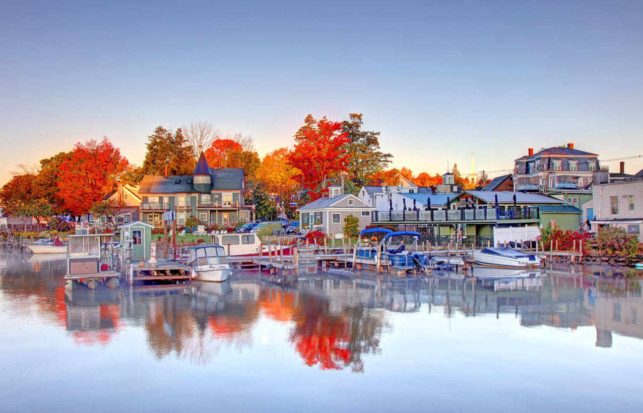 Colorful houses and boats reflected on a calm waterfront, with vibrant autumn trees in the background under a clear blue sky.