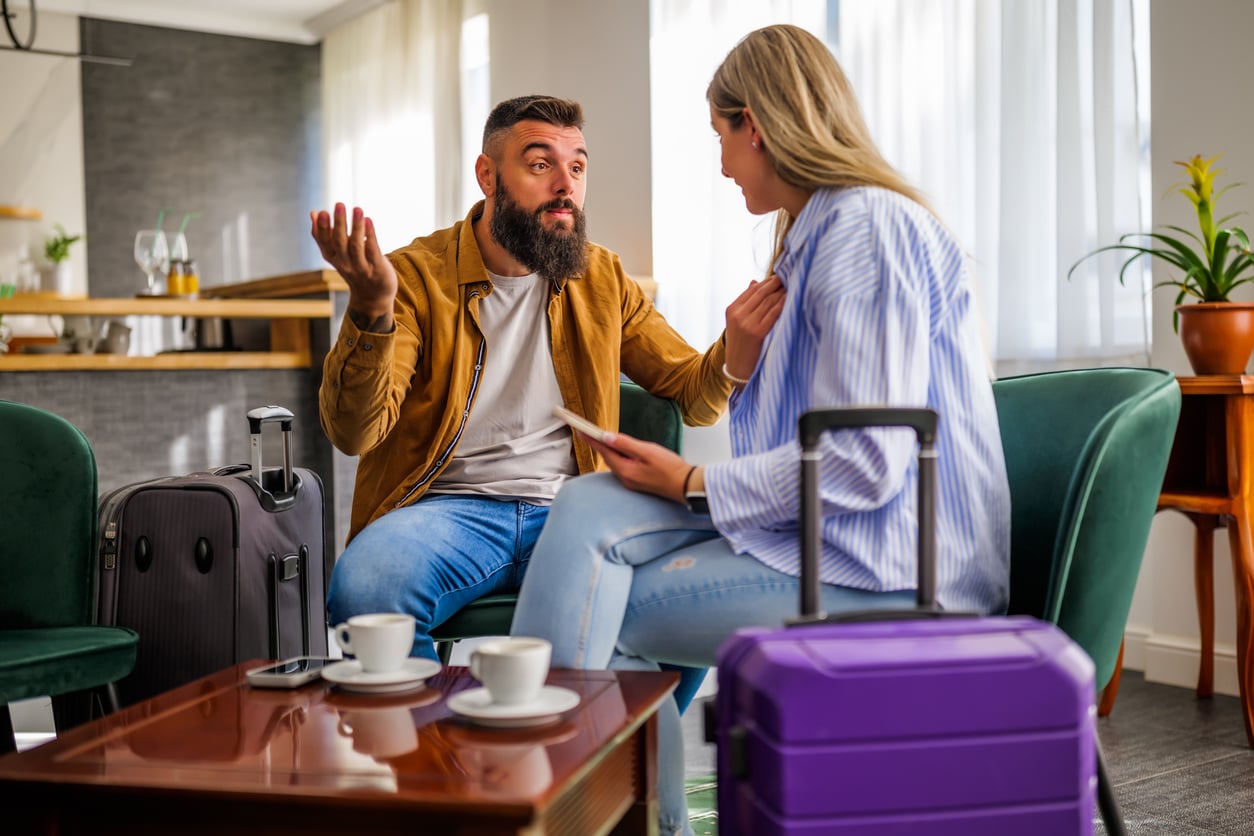 A man and woman sit in a living room with suitcases, talking seriously. The man gestures with his hands, perhaps discussing hotel booking tips, while the woman holds a piece of paper. Two coffee cups are on the table in front of them.