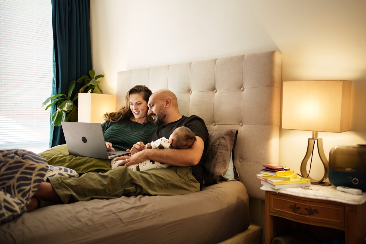A couple sits together on a bed, smiling at a laptop while one holds their baby. Warm bedside lamps, books, and a plant set the scene—reflecting how Families in America Are Having Fewer Children and cherishing cozy family moments.