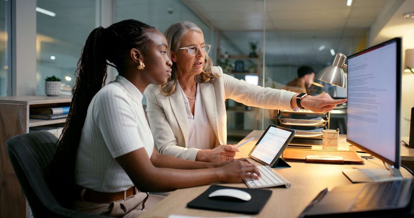Two women work together at a desk, looking at a computer screen. As they collaborate—one pointing at the monitor while the other types on a keyboard—they review documents and laptops in an office setting, reflecting how Women Are Changing Their Spending Behaviors in today's dynamic workplace.