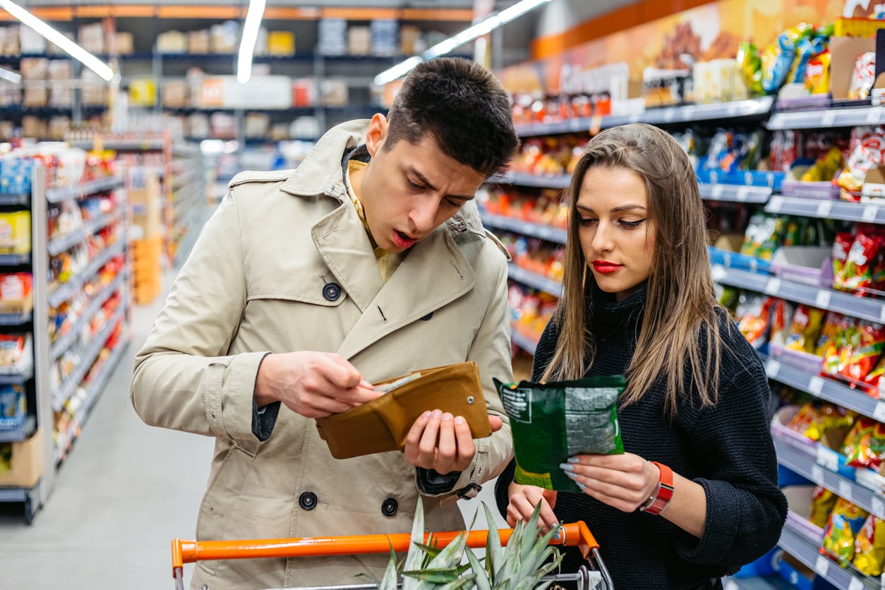 A man and woman stand beside a shopping cart in a grocery store aisle, examining a wallet and a package, appearing focused as they review prices or ingredients—perhaps considering whether these are products people no longer buy.