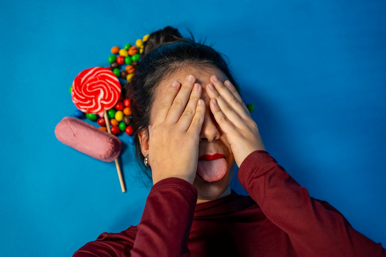 A person with red lipstick covers their eyes with their hands and sticks out their tongue, lying on a blue background with lollipops and colorful candy near their head, creating a playful scene reminiscent of fun Halloween activities.