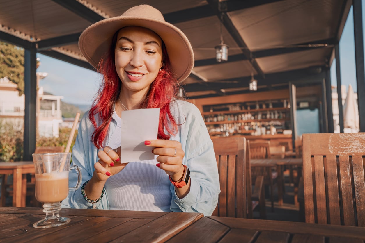 A woman with red hair and a hat smiles while looking at a piece of paper at an outdoor café, with a glass of iced coffee on the table in front of her, reflecting how women are changing their spending behaviors in everyday moments.