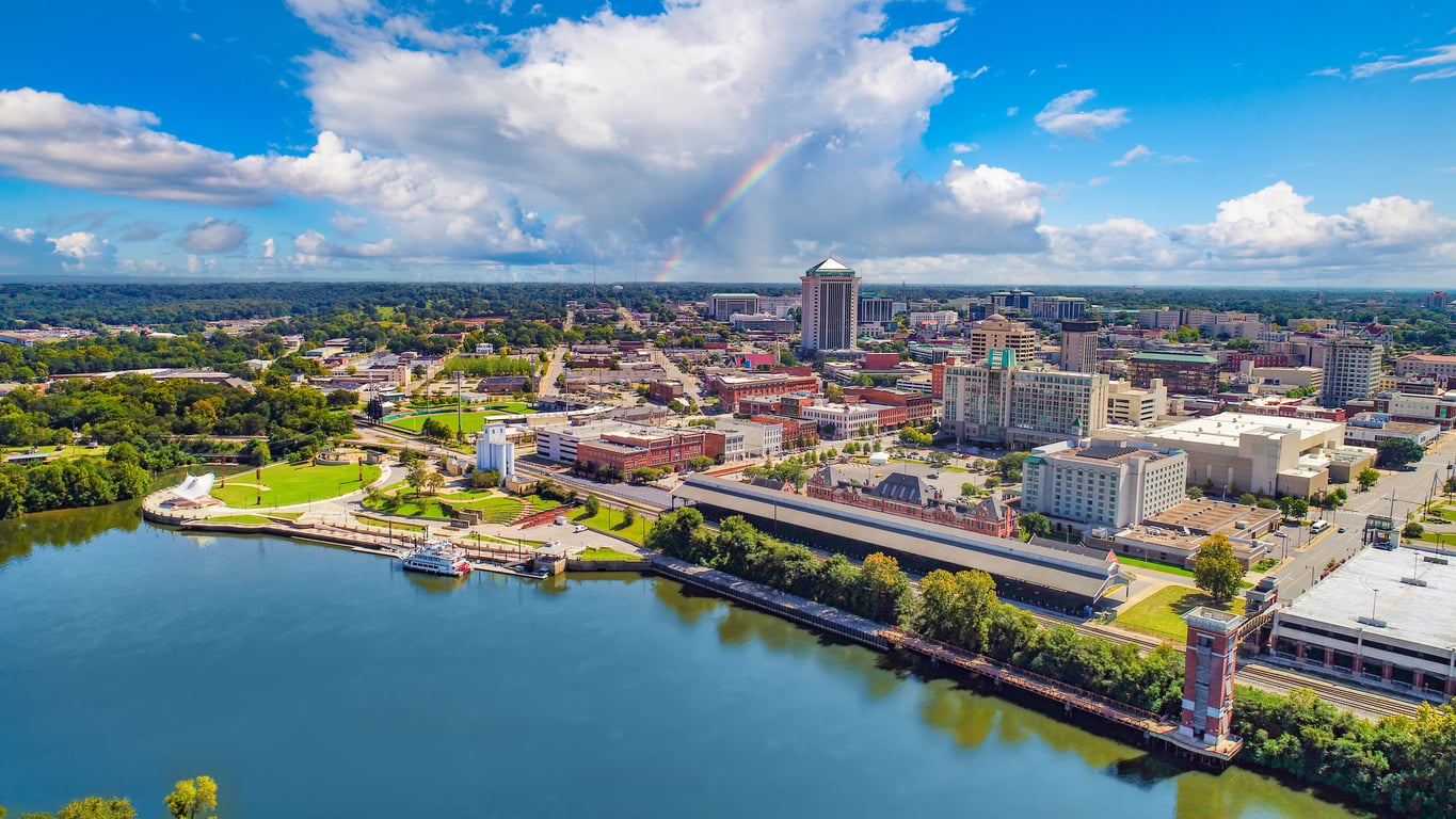 Aerial view of a travel-friendly cityscape with a river in the foreground, green trees, downtown buildings, and a partly cloudy sky with a faint rainbow visible in the distance.