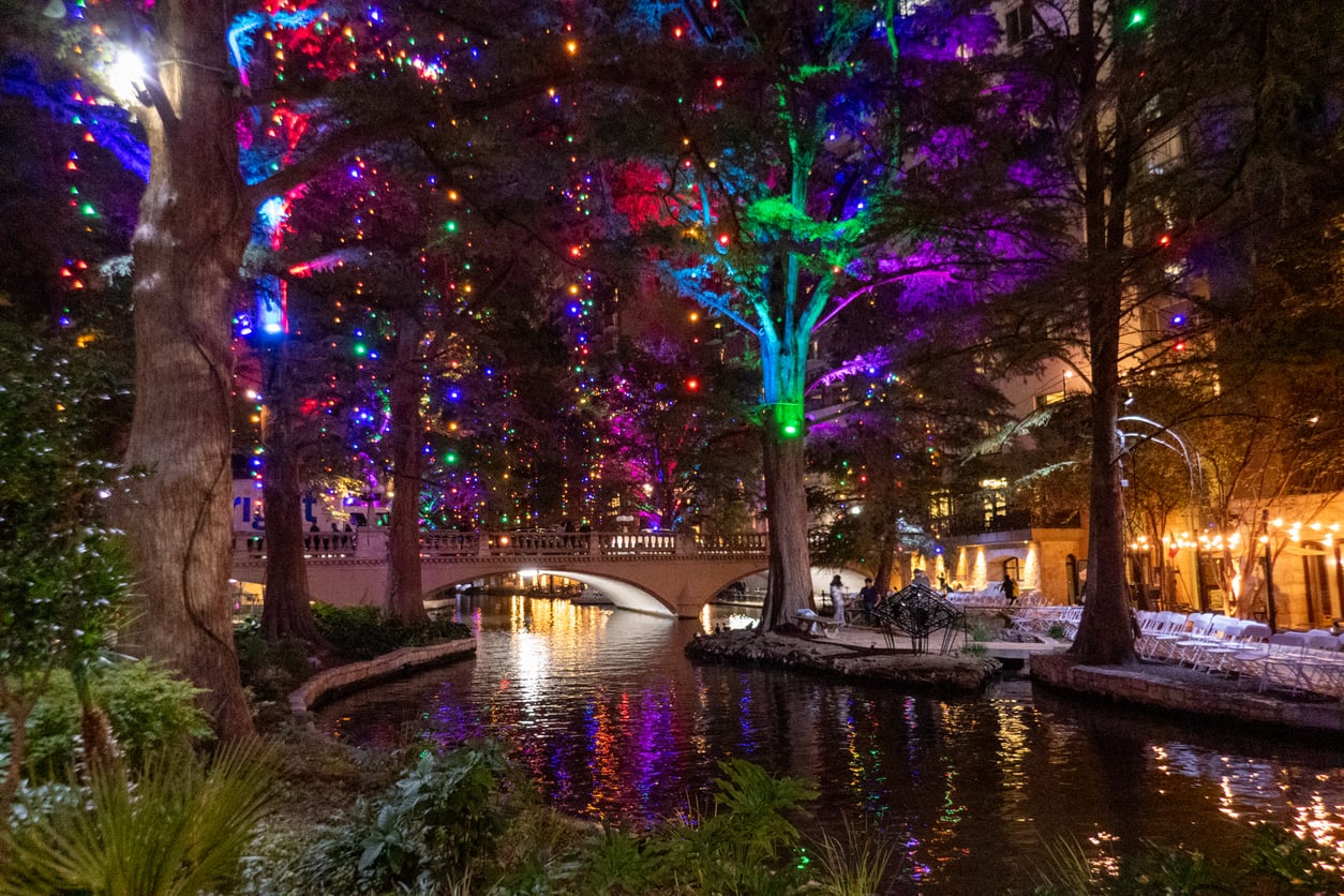 A riverbank lined with large trees decorated with colorful holiday lights, reminiscent of the festive charm found in the best Christmas towns in America, with a stone bridge crossing over the water and reflections of the lights shimmering on the surface.