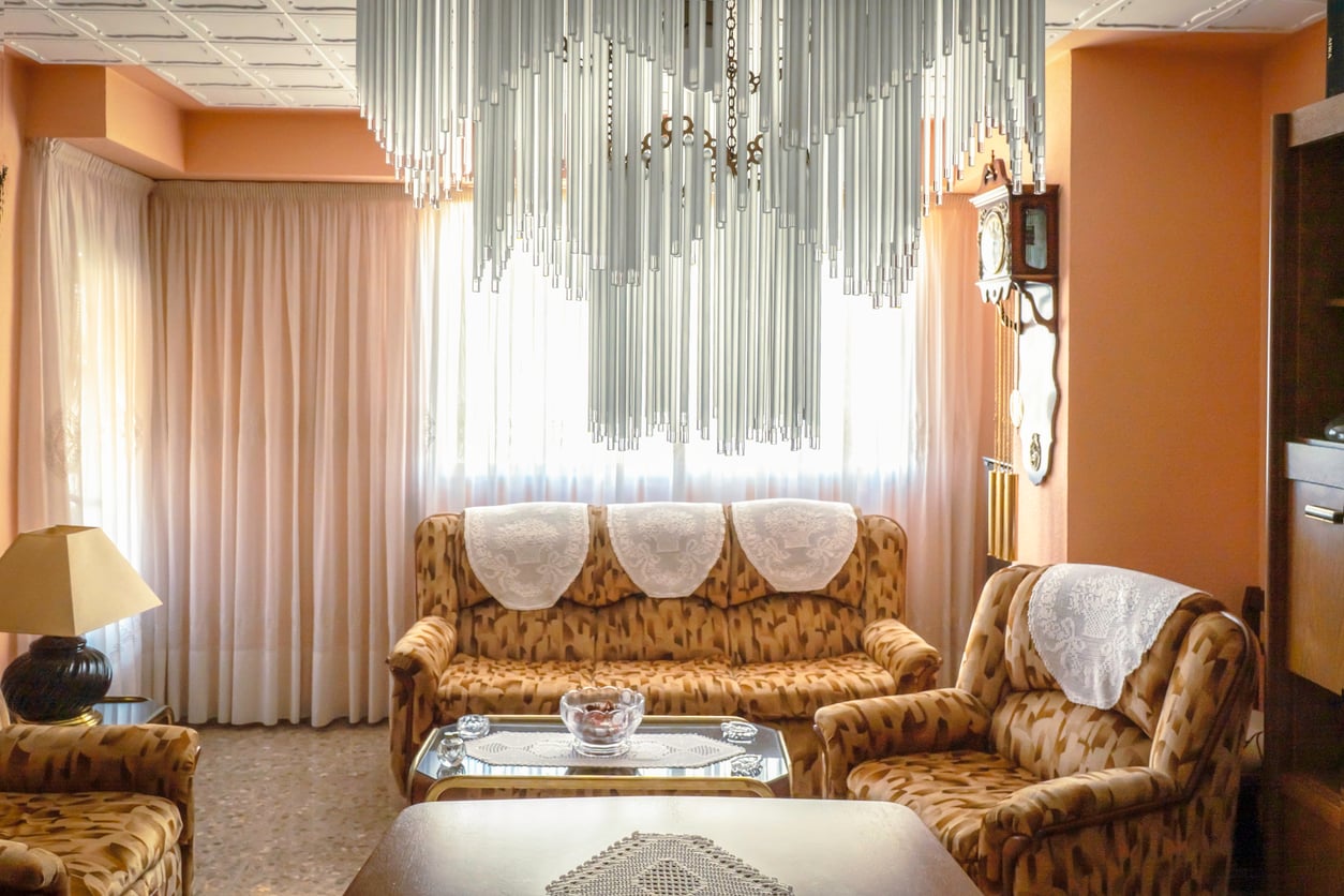 Bright living room with patterned brown sofas, lace doilies, a glass coffee table, a lamp, and a large modern chandelier—a cozy space reminiscent of the homes lovingly curated by Middle-Class Moms in the 80s and 90s. Sunlight streams through sheer curtains covering two windows, while a wall clock hangs beside the window.