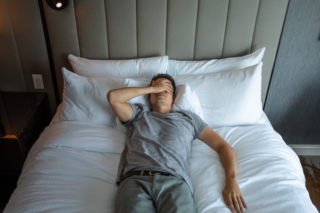 A person in a gray t-shirt and pants lies on a bed with white sheets, covering their face with one hand, appearing tired or stressed—perhaps reflecting on recent hotel changes. The room has a padded headboard and neutral decor.