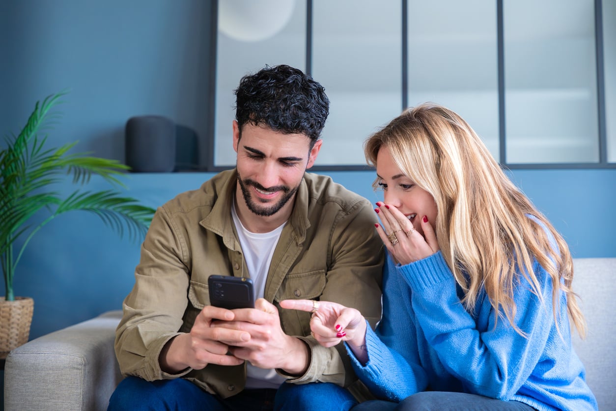 A man and woman sit on a couch, smiling and looking at a smartphone. The woman covers her mouth with one hand and points at the phone with the other. A plant and blue wall are in the background.
