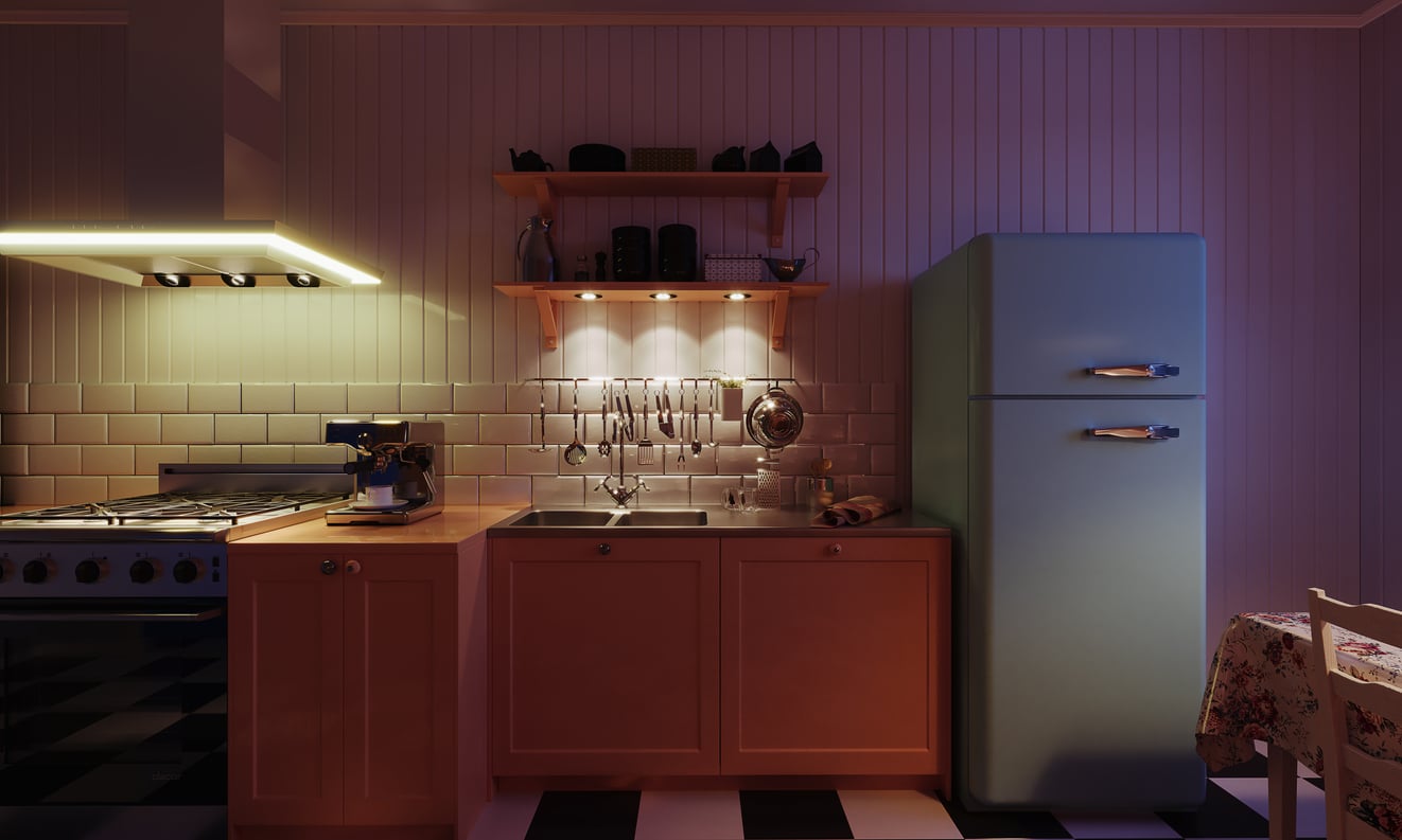 A cozy kitchen with warm lighting, a blue retro refrigerator, wooden cabinets, a sink, utensils hanging on a tiled backsplash, shelves with dishes, and a stove on the left. A floral tablecloth is partly visible on the right.