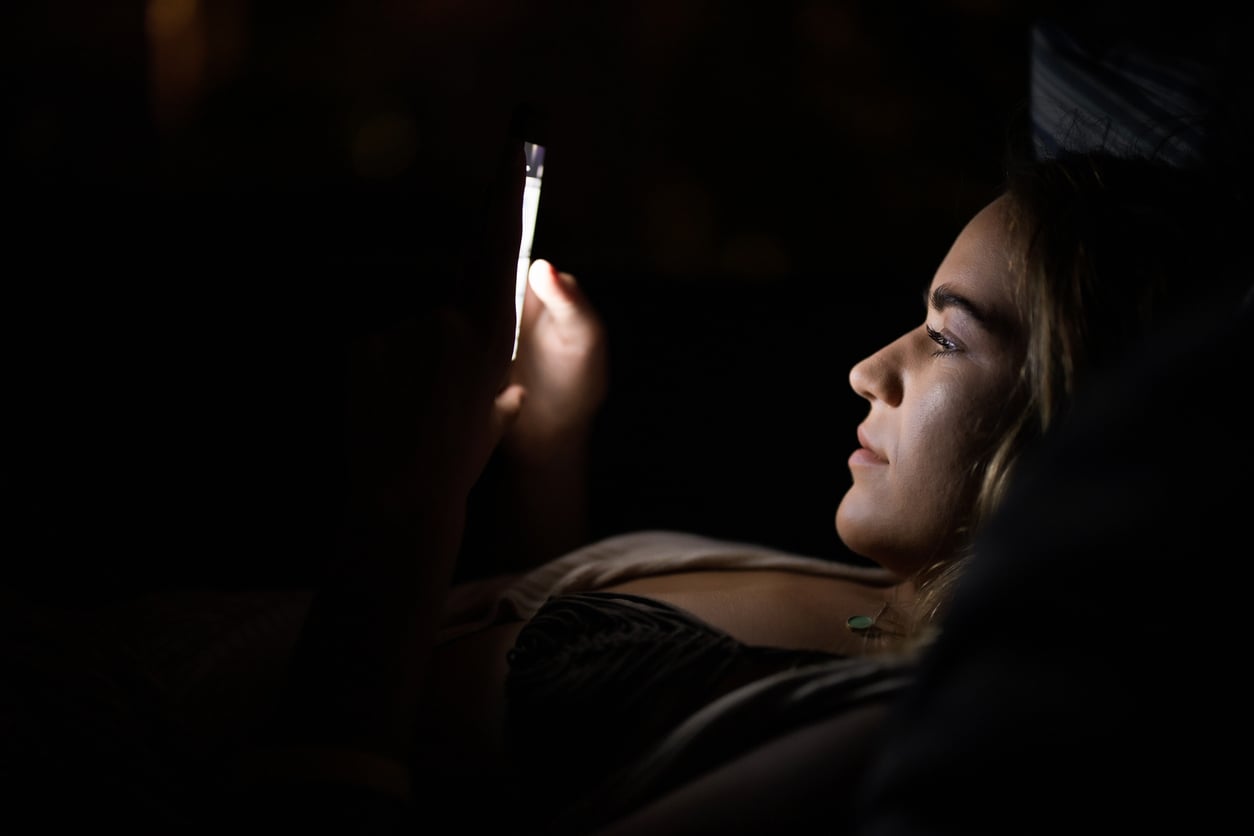A person lies in bed in a dark room, illuminated by the light from a smartphone they are holding and looking at closely.
