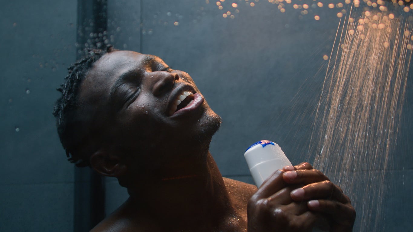 A man with short, twisted hair smiles and closes his eyes while standing under a shower, holding a white bottle, with water streaming over him in a dark tiled bathroom.
